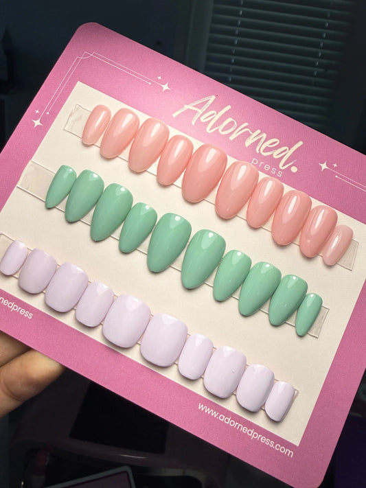 3 sets of press ons in different shapes and colors - pink, pistachio green, and lavender - on a backing card that says Adorned Press