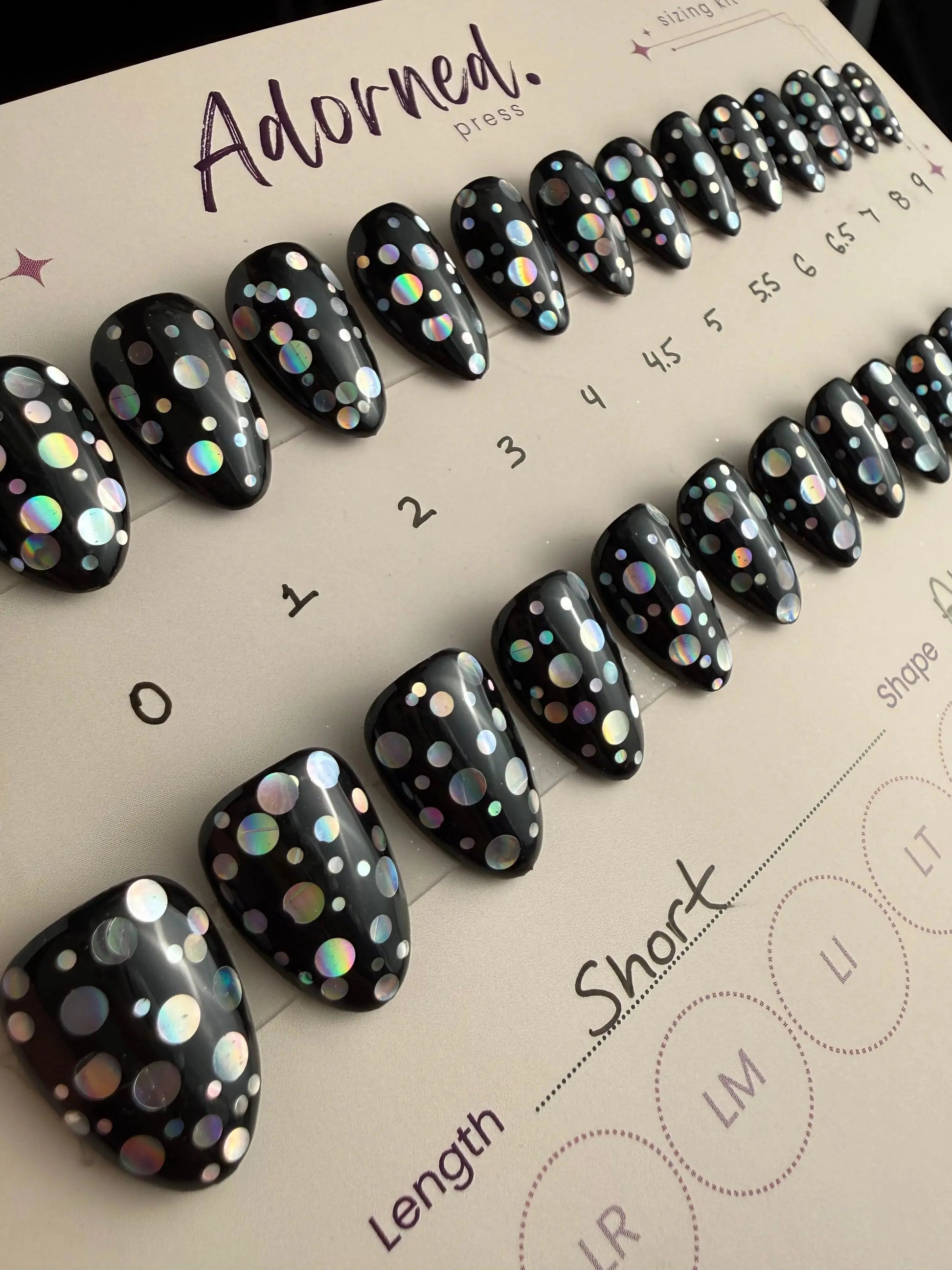 Black nail art with multicolored dots on a 'Adorned' press display.